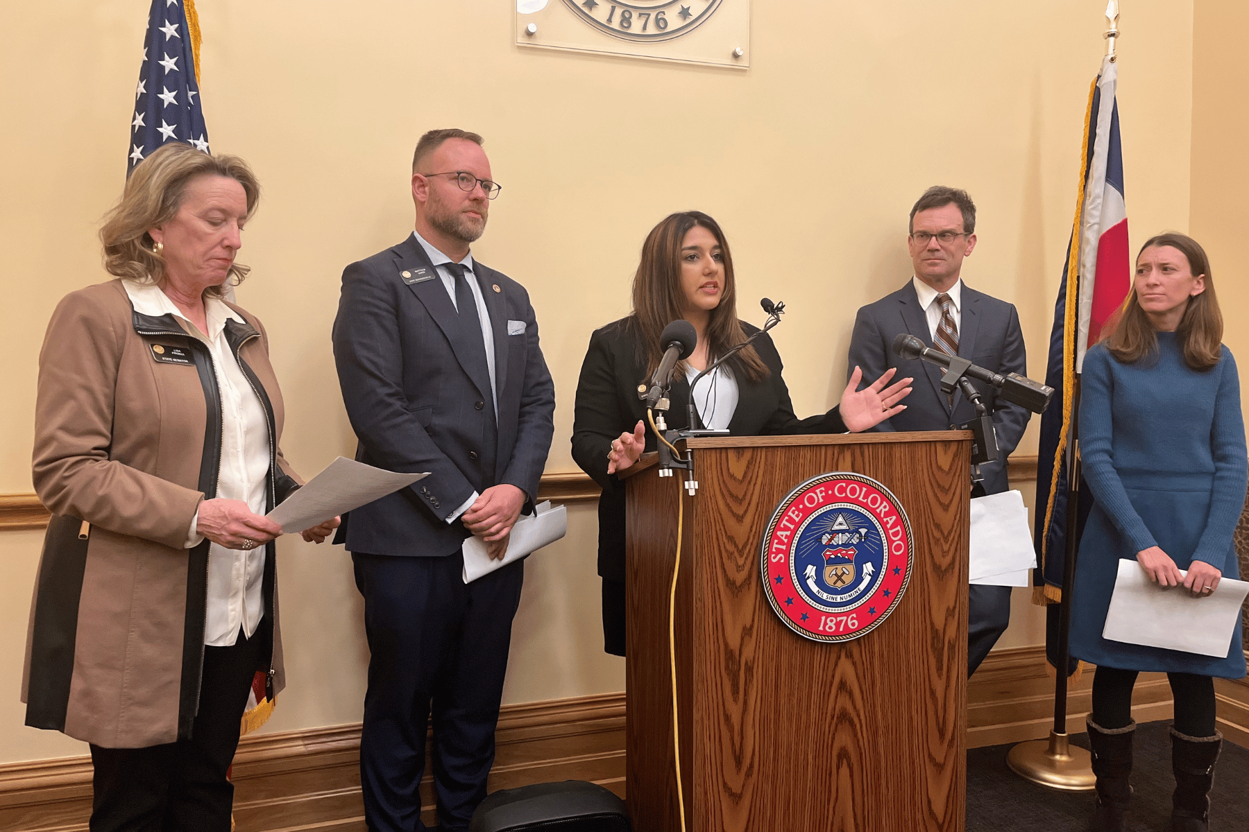 Democratic Rep. Yara Zokaie stands at a podium, flanked by two more lawmakers and two advocates. The flags of Colorado and the U.S. are on stands behind them.