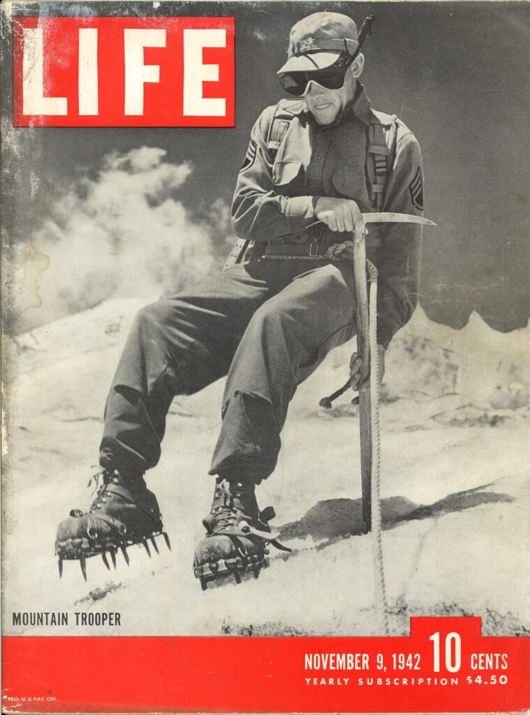 Photo shows a black and white image of a member of the 10th Mountain Division on a snowy mountain wearing spiked boots and holding a pick axe to use on ice.