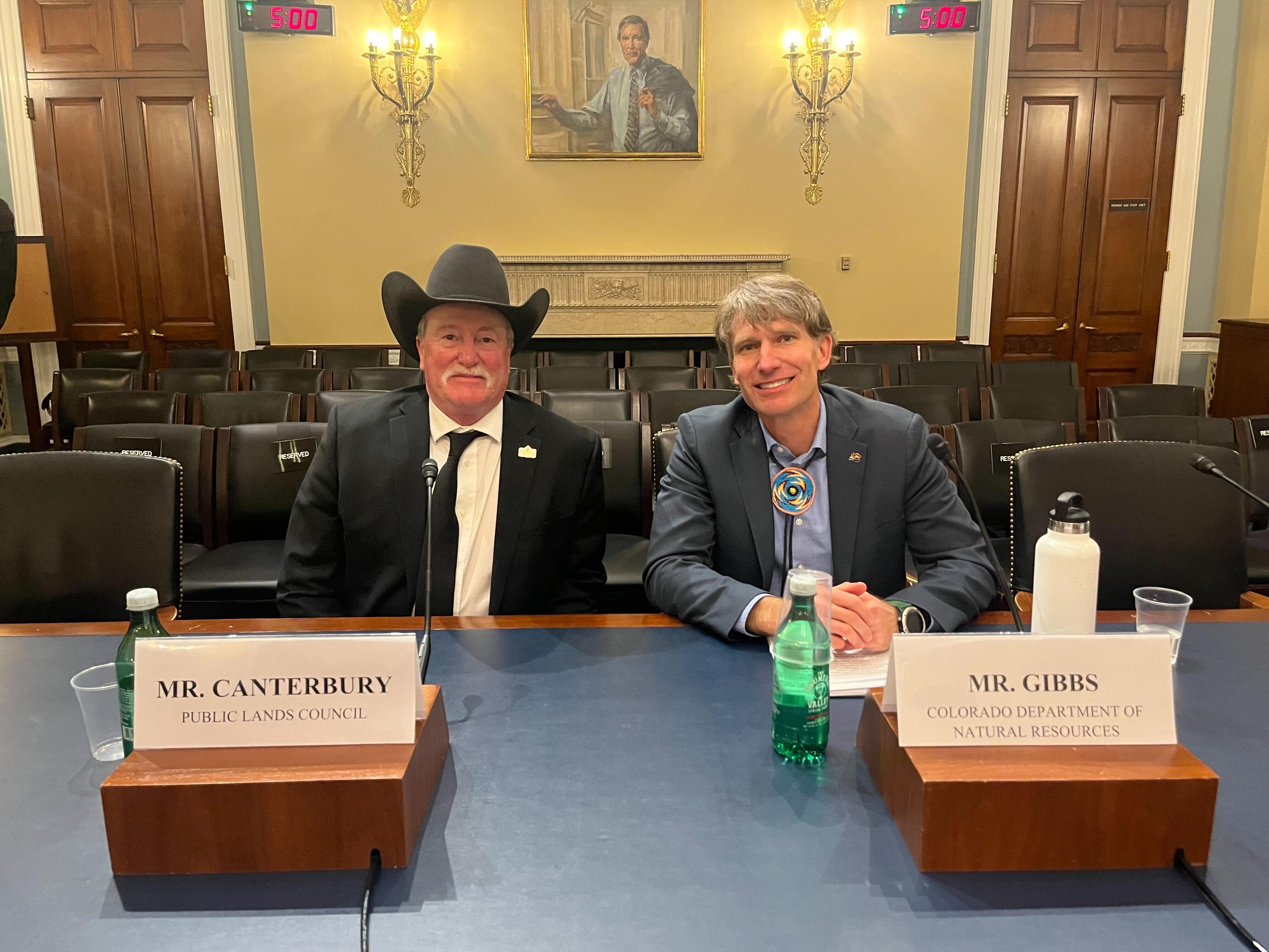 Two men, Canterbury and Gibbs, sit side by side in the hearing room.