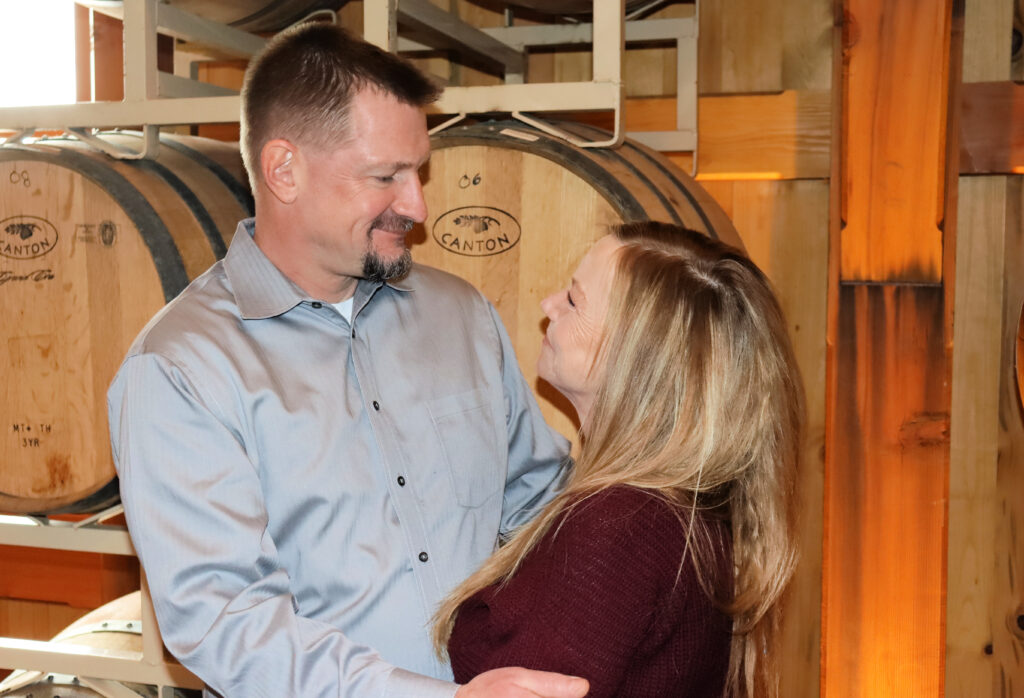 A couple smile at each other in front of rustic wine barrels.