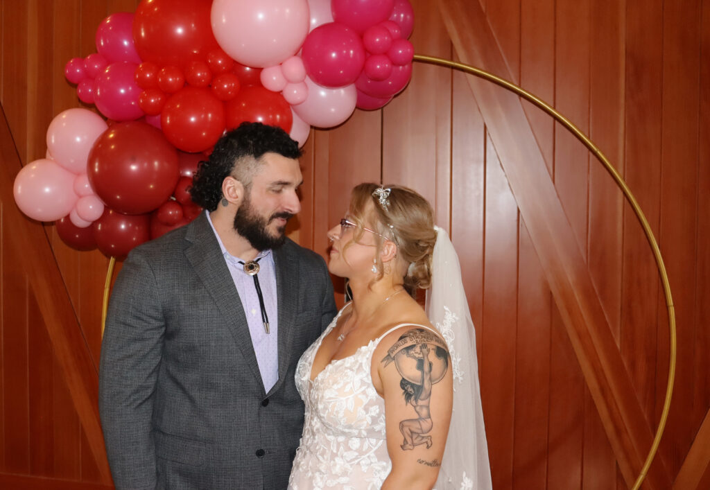 A young couple pose for a photo under pink, red, and white balloons.