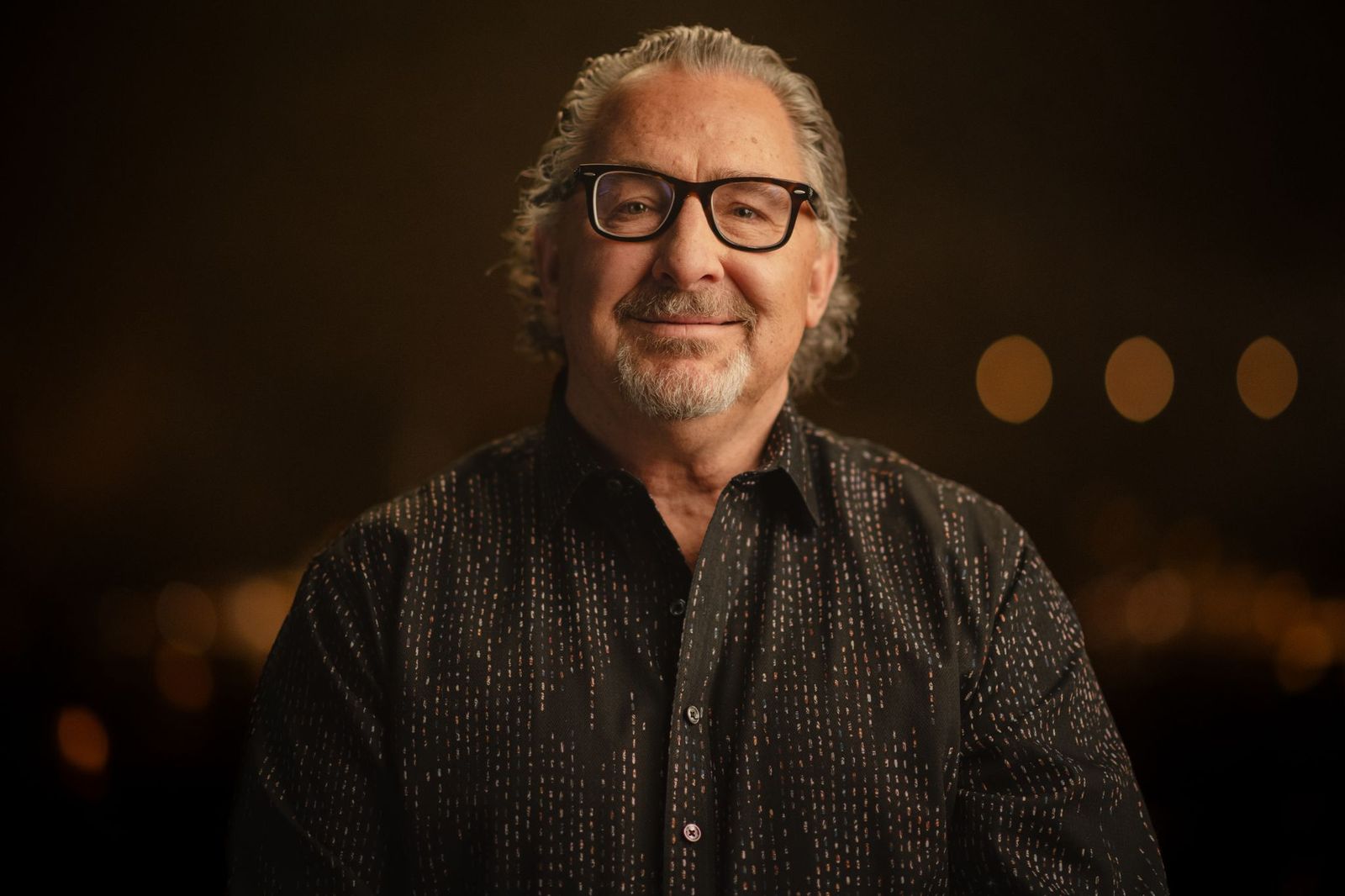 A man in a black button-up shirt, gray hair and black glasses looks at the camera