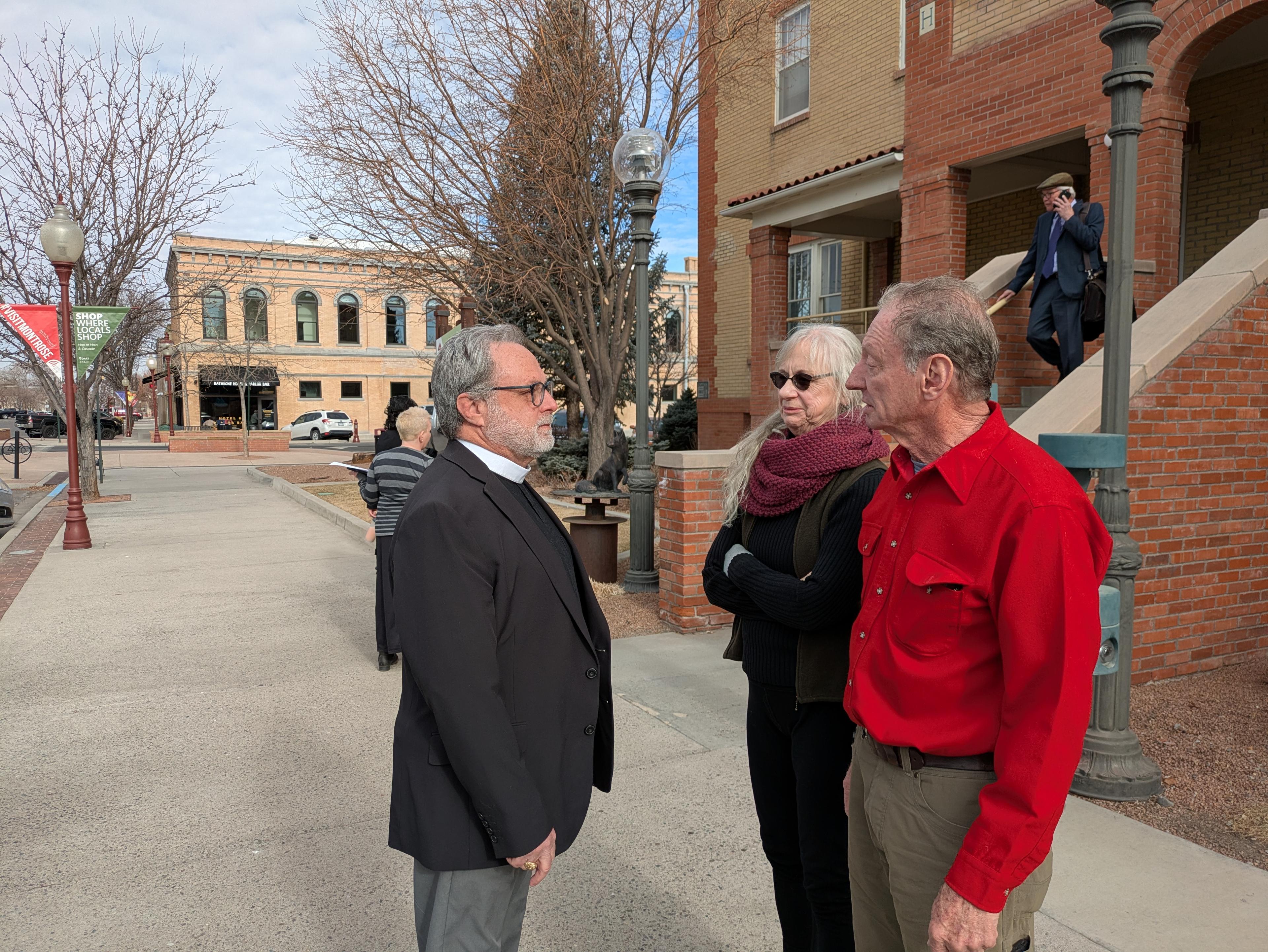 Kevin Young, wearing a charcoal jacket and a clerical collar, speaks with supporters outside of a two-story historic brick building where Montrose Municipal Court hearings take place.