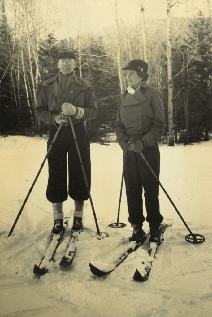 A black and white photo shows Charles Minot “Minnie” Dole and his wife Jane on skis with ski poles in front of trees on a snowy slope.