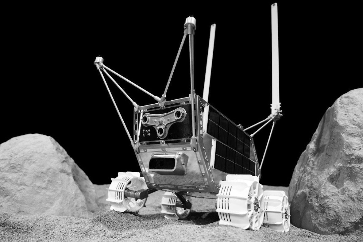 a silver box-shaped robotic lunar rover with antennas sticking up rolls around the lunar surface near two large Moon rocks.