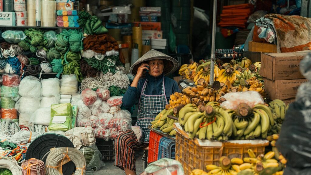 An Vietnamese woman in a conical hat sits behind a table of bananas in a busy street market scene.