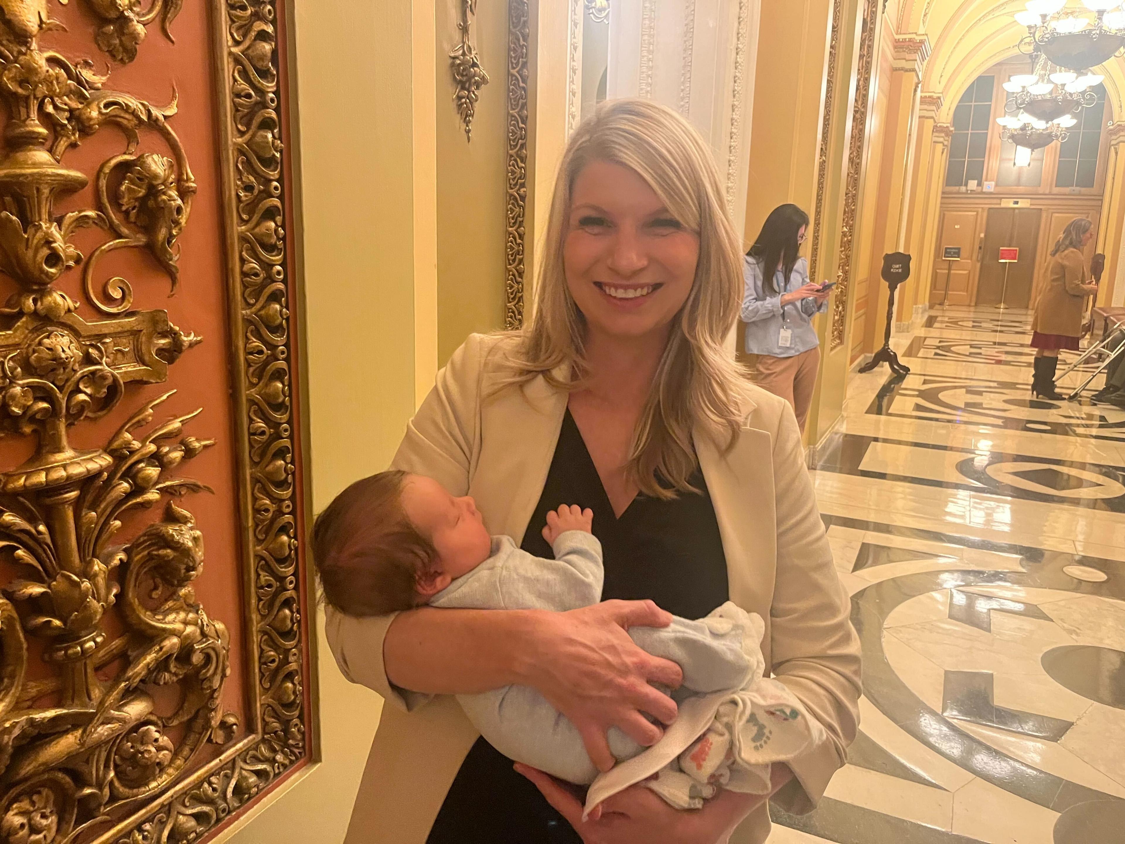 Rep. Brittany Pettersen holds a sleeping baby in her arms while standing in a capitol hallway.