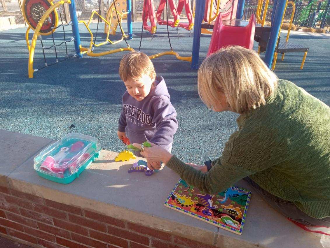 A boy plays with block puzzle pieces at a play ground as a woman watches over him..