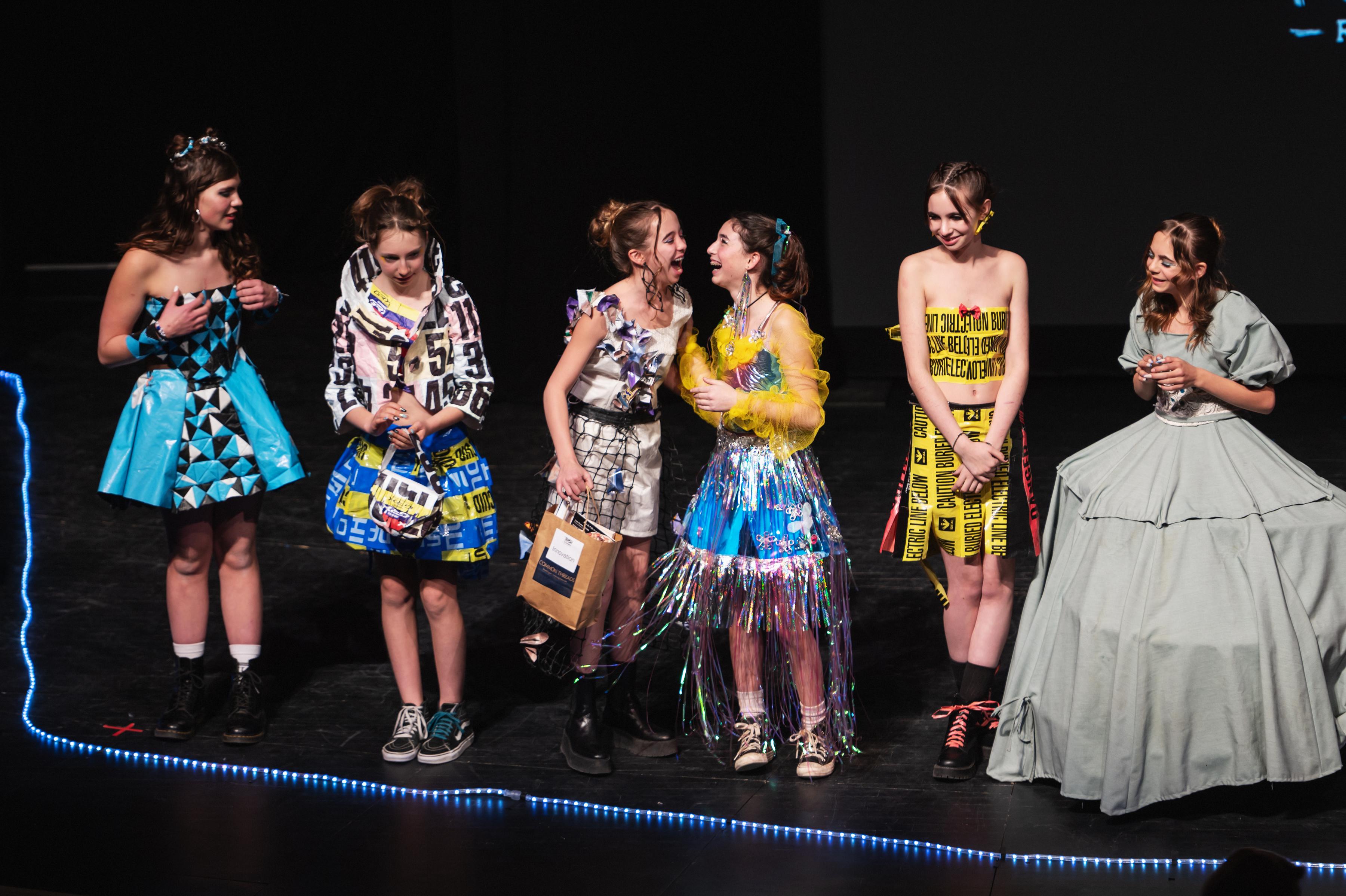 Students wearing outfits they designed from trash are seen in a line on stage.