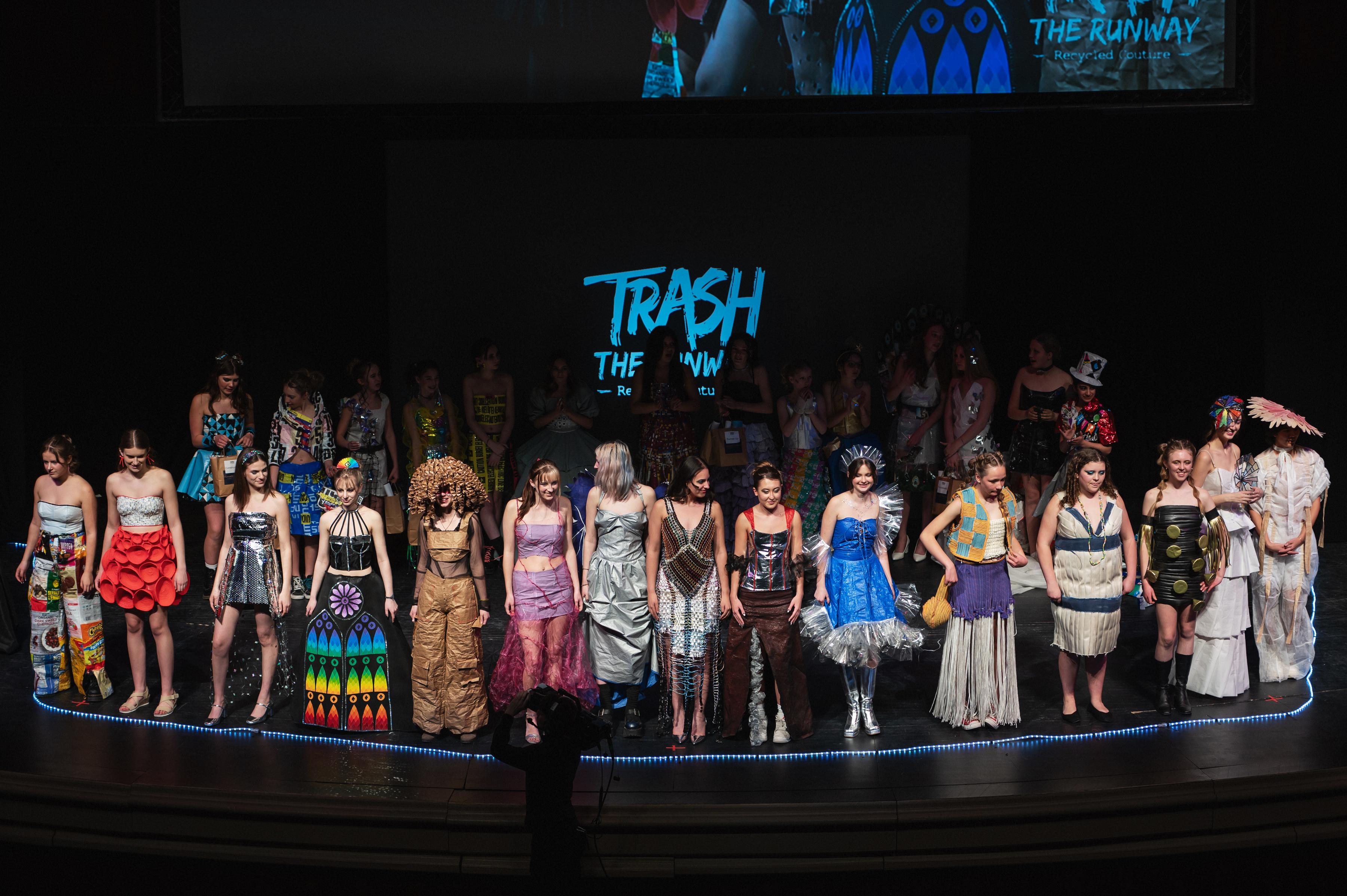 Students wearing outfits they designed from trash are seen in a line on stage.