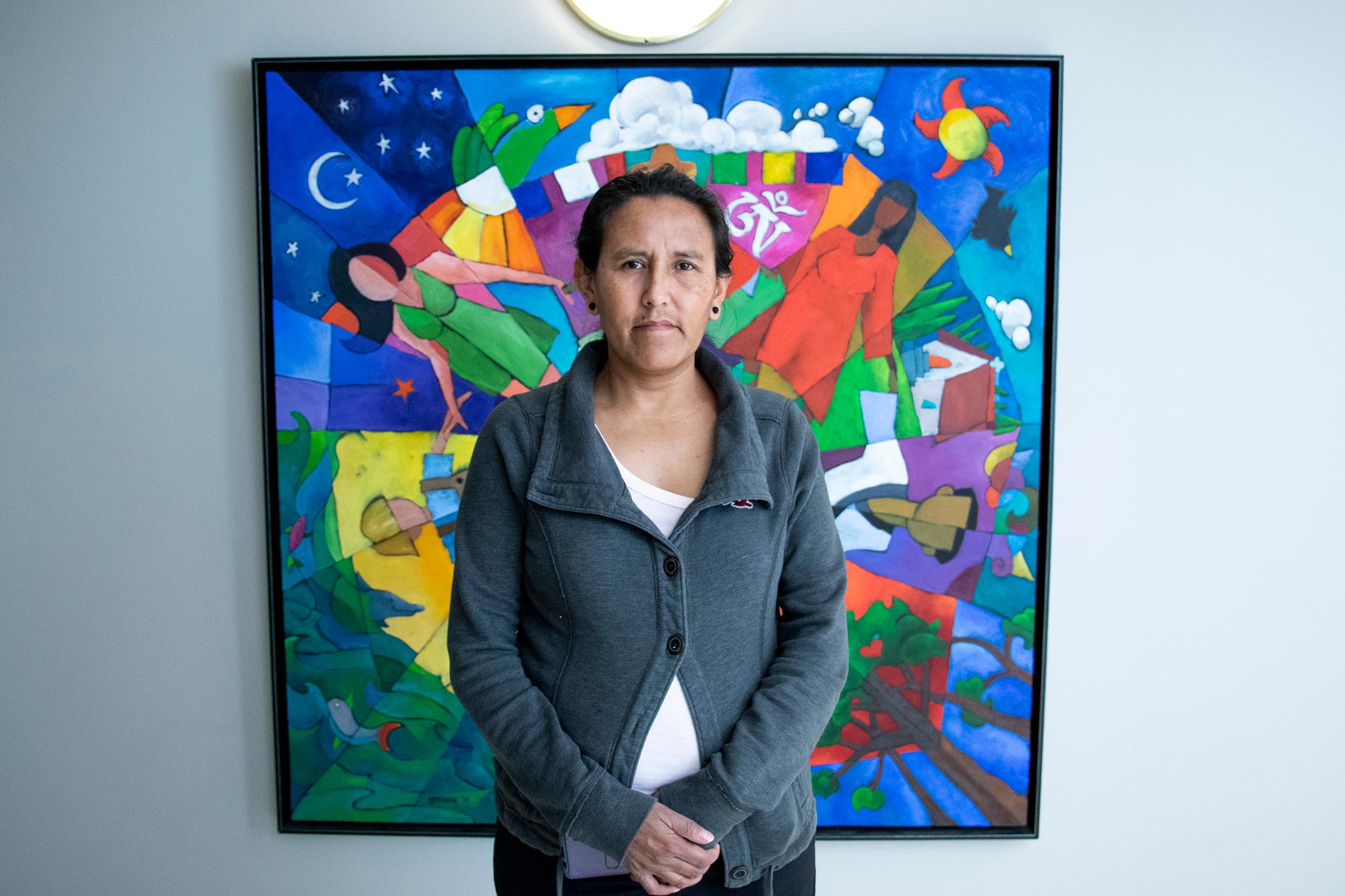 A woman in a gray sweater stands in front of a colorful painting with her hands folded.