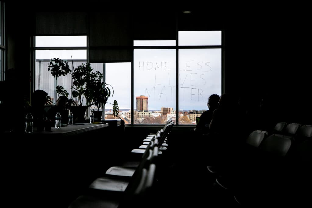 A dark room with two people sitting near a window that reads, "Homeless lives matter" in all caps.