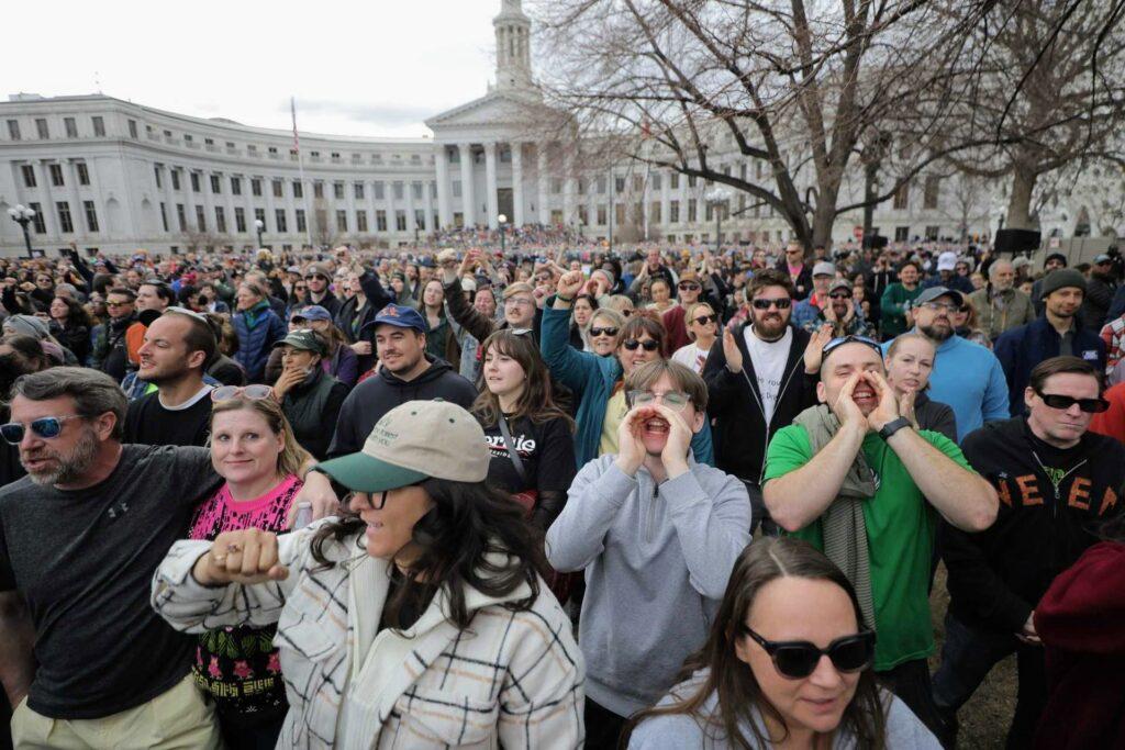 A large crowd of people cheer and raise their fists in front of the Denver City and County Building.