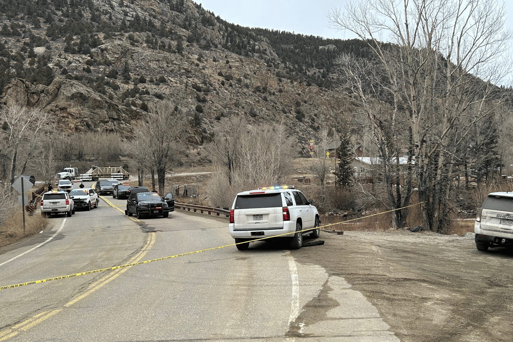 Nine white and black police and sheriff's vehicles with flashing lights are parked on a small bridge over a river near trees and foothills.