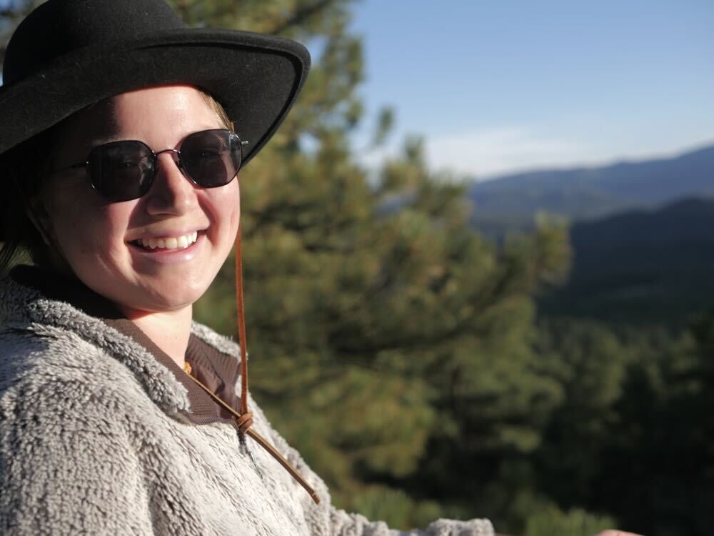 A woman in sunglasses and a hat sits on a hill with a green tree in the background.