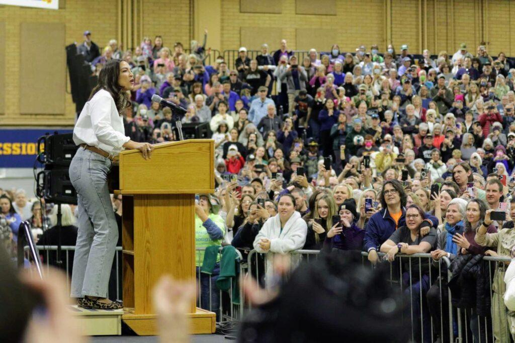 Alexandria Ocasio-Cortez speaks from a podium at the Bank of Colorado Arena in Greeley while eager supporters watch. Many have their phones out to record the moment.