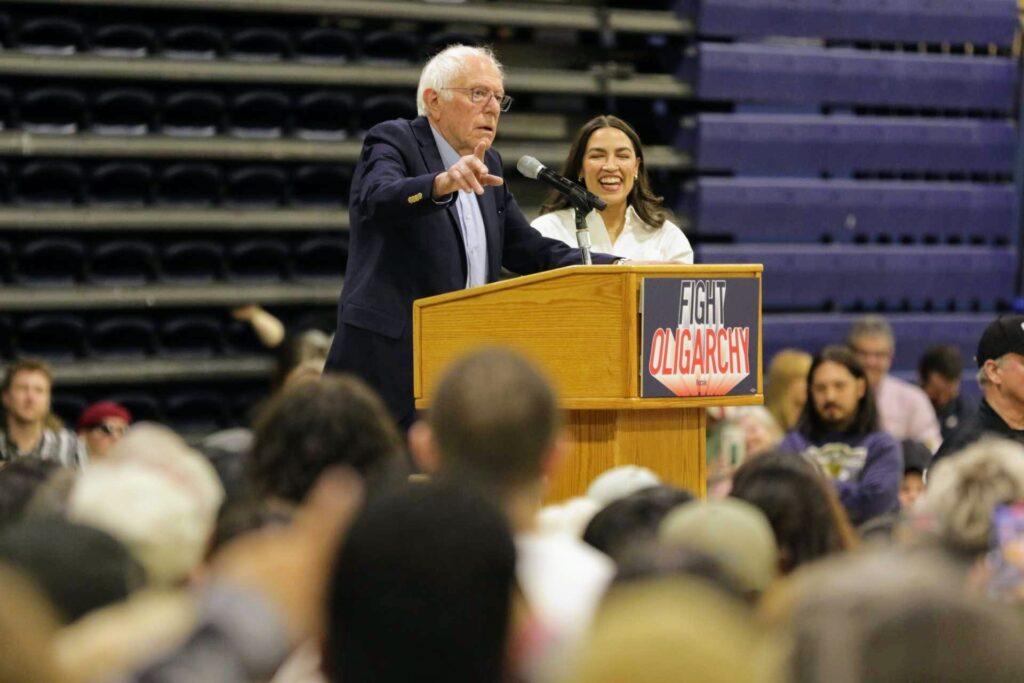 Bernie Sanders and Alexandria Ocasio-Cortez stand together at a podium reading &quot;Fight Oligarchy&quot; from a podium that reads &quot;Fight Oligarchy,&quot; surrounded by a large crowd.