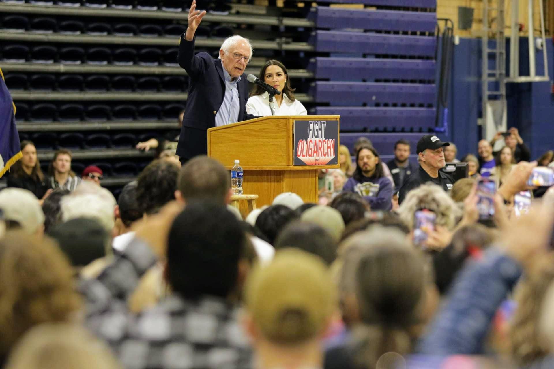 Bernie Sanders and Alexandria Ocasio-Cortez stand together at a podium reading "Fight Oligarchy" from a podium that reads "Fight Oligarchy," surrounded by a large crowd.