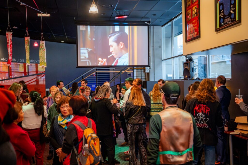 People gathered in the lobby of the SIE FilmCenter for the 2024 Dragon Boat Film Festival. Asian decorations hang from the ceiling and a movie is projected on the back wall.