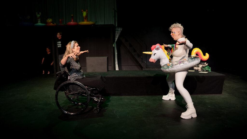 Two actors, one in a wheelchair, one dressed as a unicorn, face each other with hands out as if they're about to play fight.