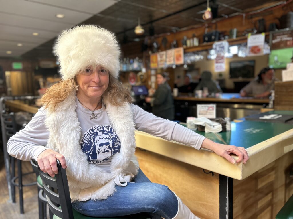 Stephanie Andelman sits at a bar in Nederland, Colorado wearing a &quot;Frozen Dead Guy Day&quot; T-shirt and a white faux fur hat and vest.