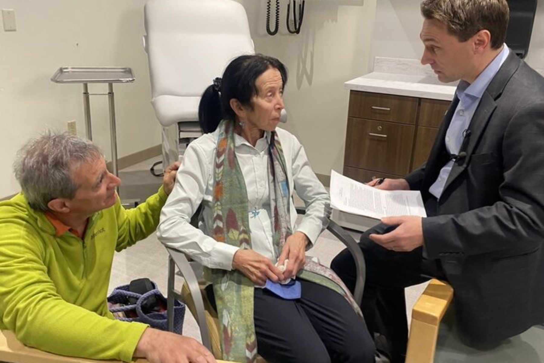 From left, a man in a green shirt touches a seated woman's arm as they both look at a man holding a sheet of paper. They are all in a doctor's office exam room.