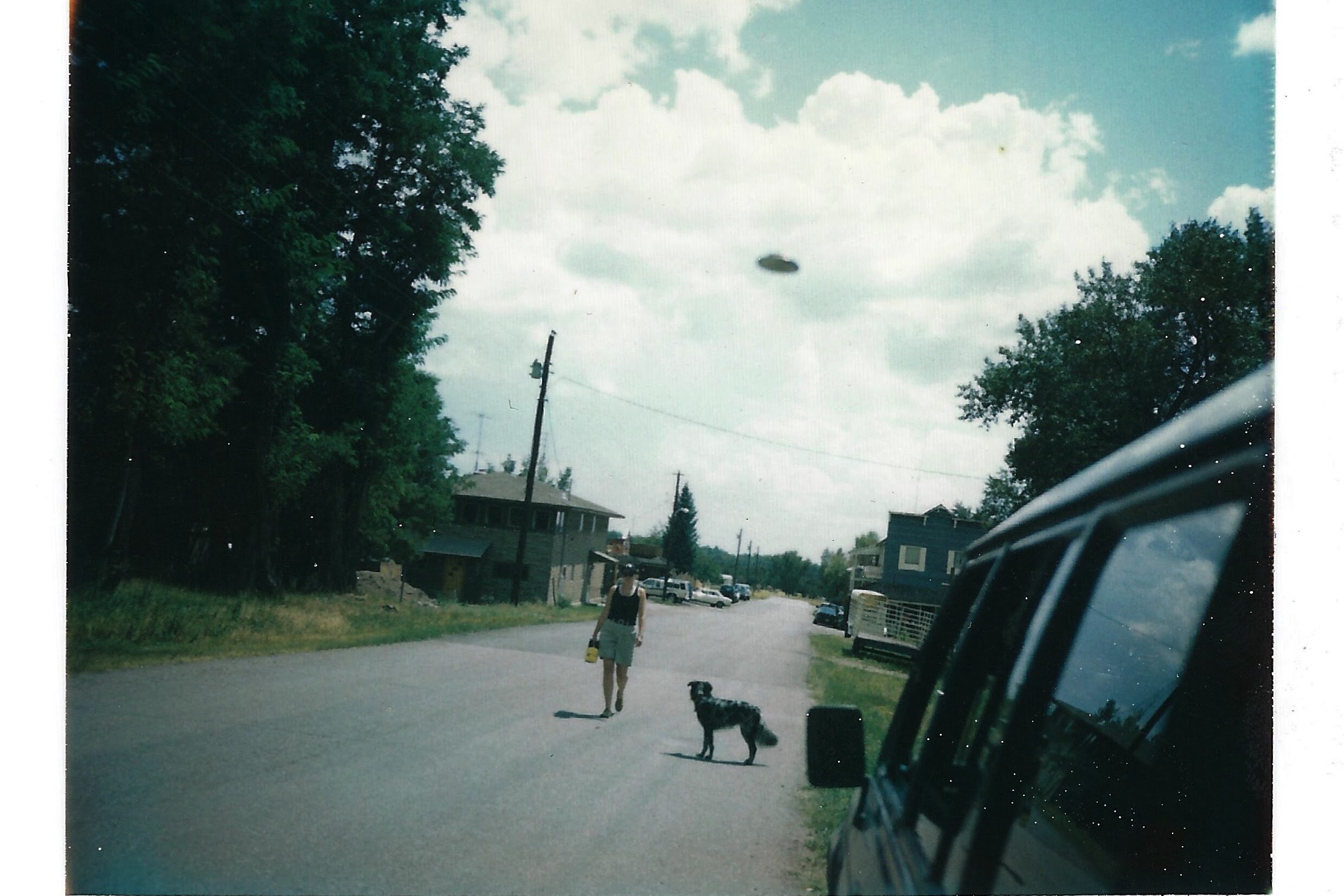 A Polaroid photograph of a woman and a dog walking down the street with a UFO in the sky behind them.