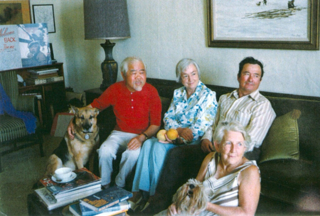 Three people sit on a sofa, one petting a dog, and one on the floor with a dog