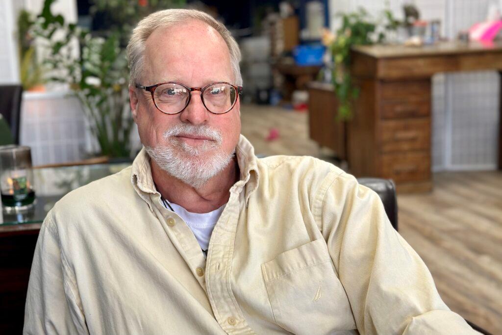 A man with a white beard, glasses and a yellow button-up shirt sits for a portrait.