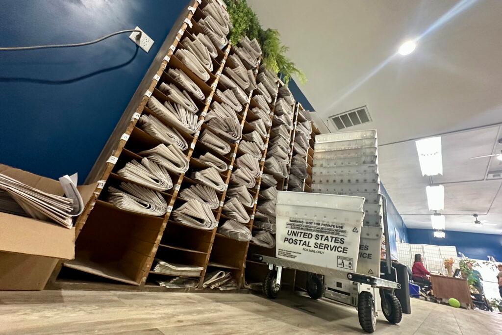 A set of shelves is packed with folded newspapers. Boxes from the US Postal Service are on a dolly in front of the shelves.