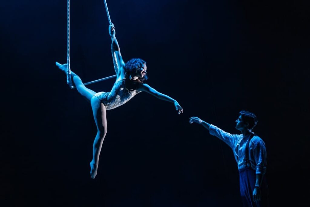 A female trapeze artist reaches her hand for a male dancer standing below her.