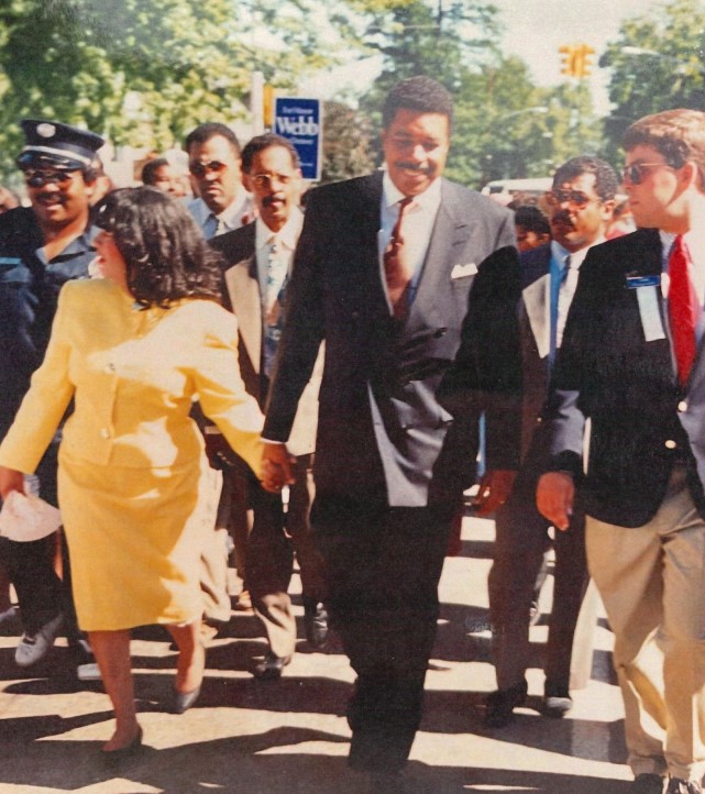 Wilma Webb is seen walking in a yellow jacket and skirt on the left side of the photo holding hands with Wellington Webb in a dark suit as they walk in the annual Marade on MLK Day in Denver.