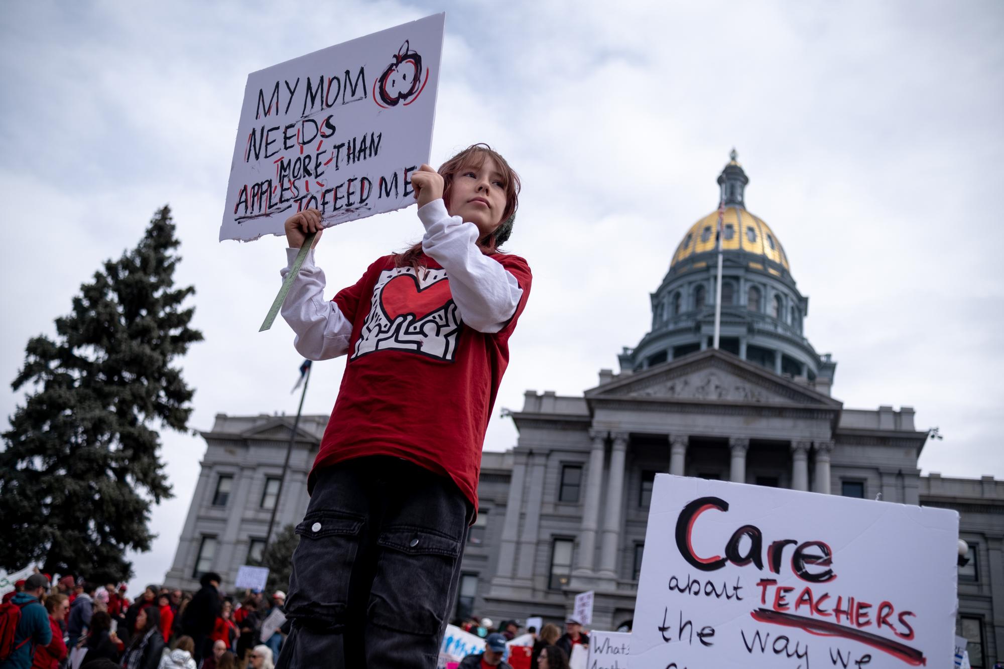 A girl holds up a sign that reads "My mom needs more than Apples to feed me." The golden Capitol dome is seen in the background.